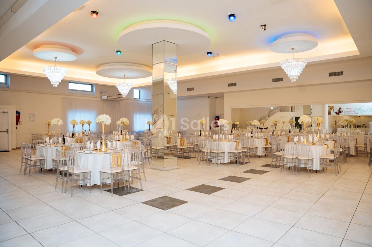 Salle de réception lumineuse avec tables décorées de fleurs blanches, chaises transparentes et lustres suspendus.