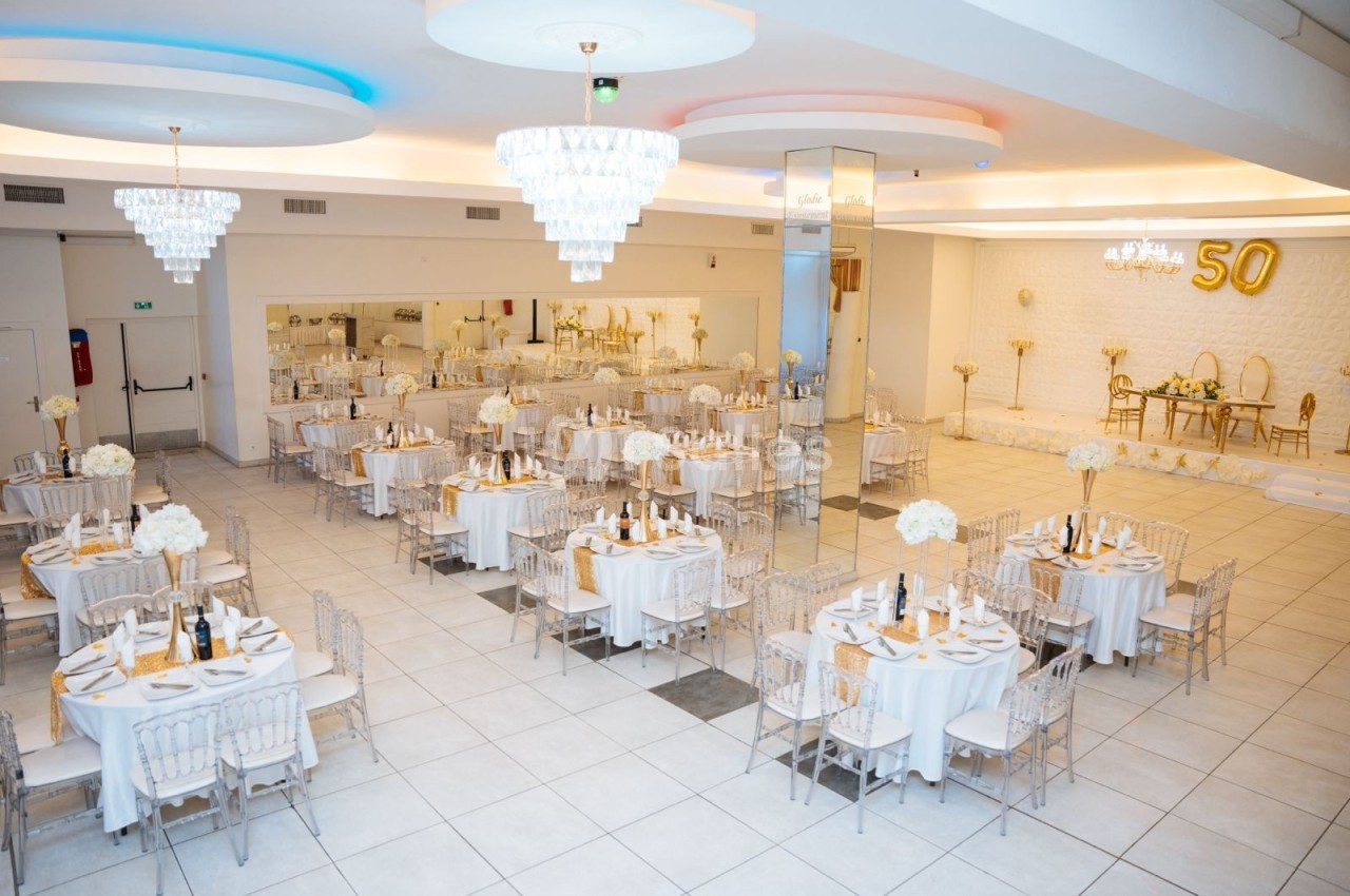 Salle de réception élégante avec tables rondes dressées, décorations blanches et dorées, et un chiffre 50 en ballon.