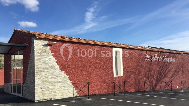 Bâtiment avec façade rouge et mur en pierre, parking asphalté à l'avant, ciel bleu en arrière-plan.