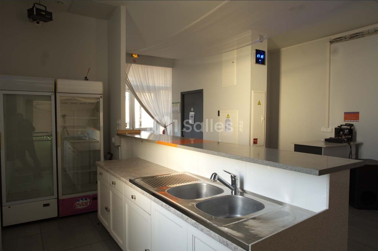 Cuisine lumineuse avec un évier double, un comptoir, un réfrigérateur vitré et une machine à café en arrière-plan.