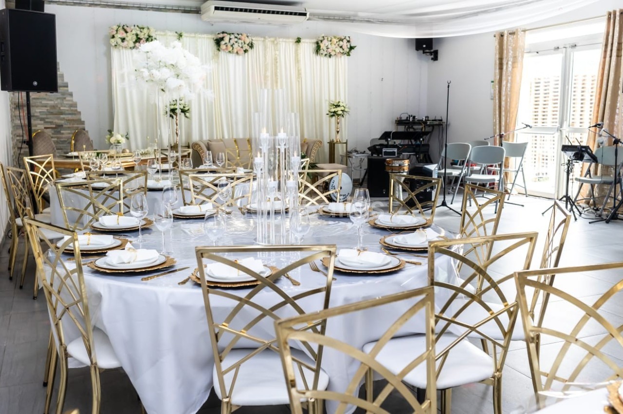 Salle de réception décorée avec des tables rondes, nappes blanches, chaises dorées et arrangements floraux élégants.