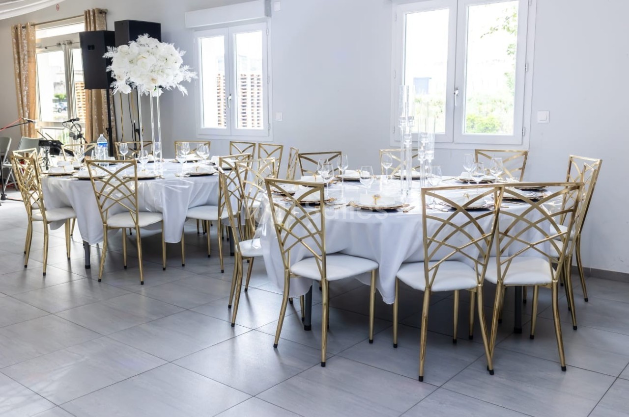 Salle lumineuse avec des tables rondes dressées, nappes blanches, chaises dorées et centre de table floral.