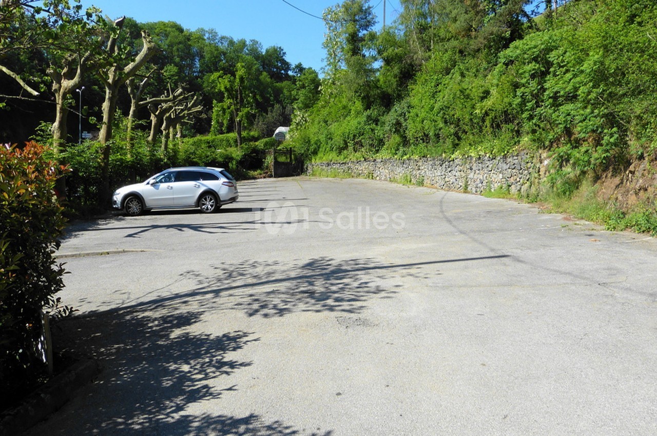 Parking extérieur vide avec un seul véhicule blanc, entouré de végétation et d'un mur en pierre.