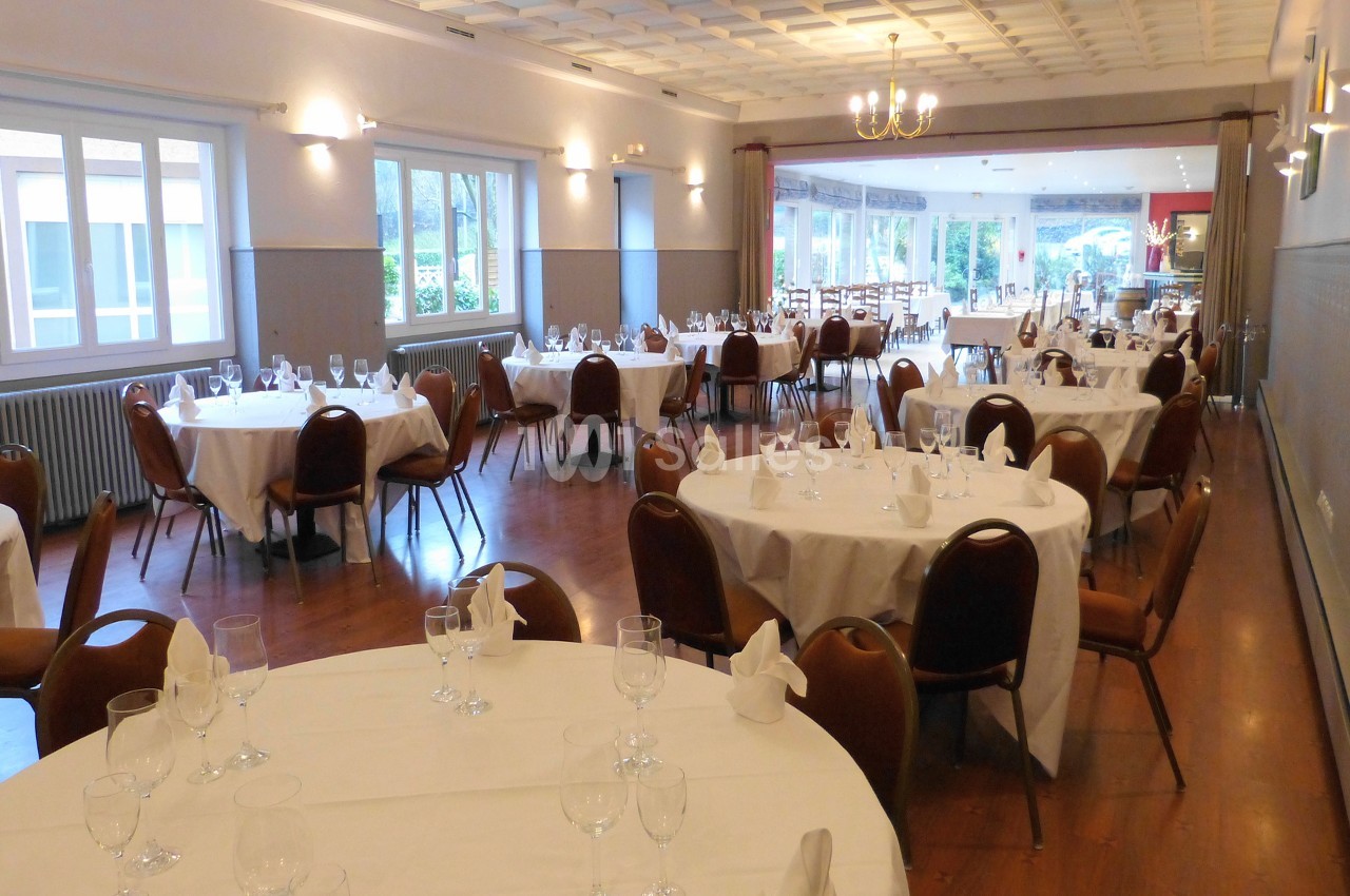 Salle de réception lumineuse avec tables rondes dressées, chaises en bois et grandes fenêtres donnant sur l'extérieur.