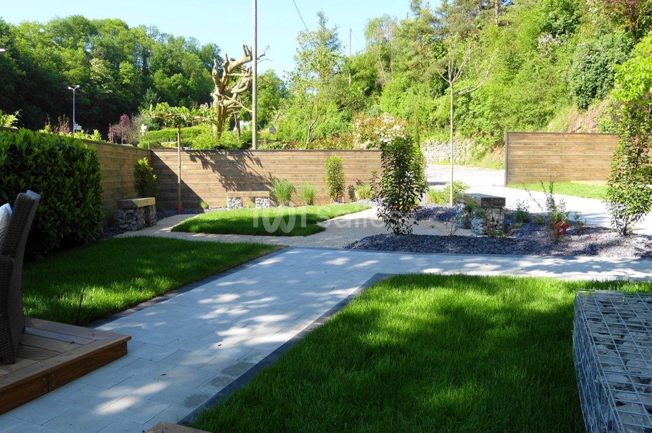 Jardin aménagé avec pelouse, allées pavées, parterres de plantes et clôtures en bois, entouré de verdure.