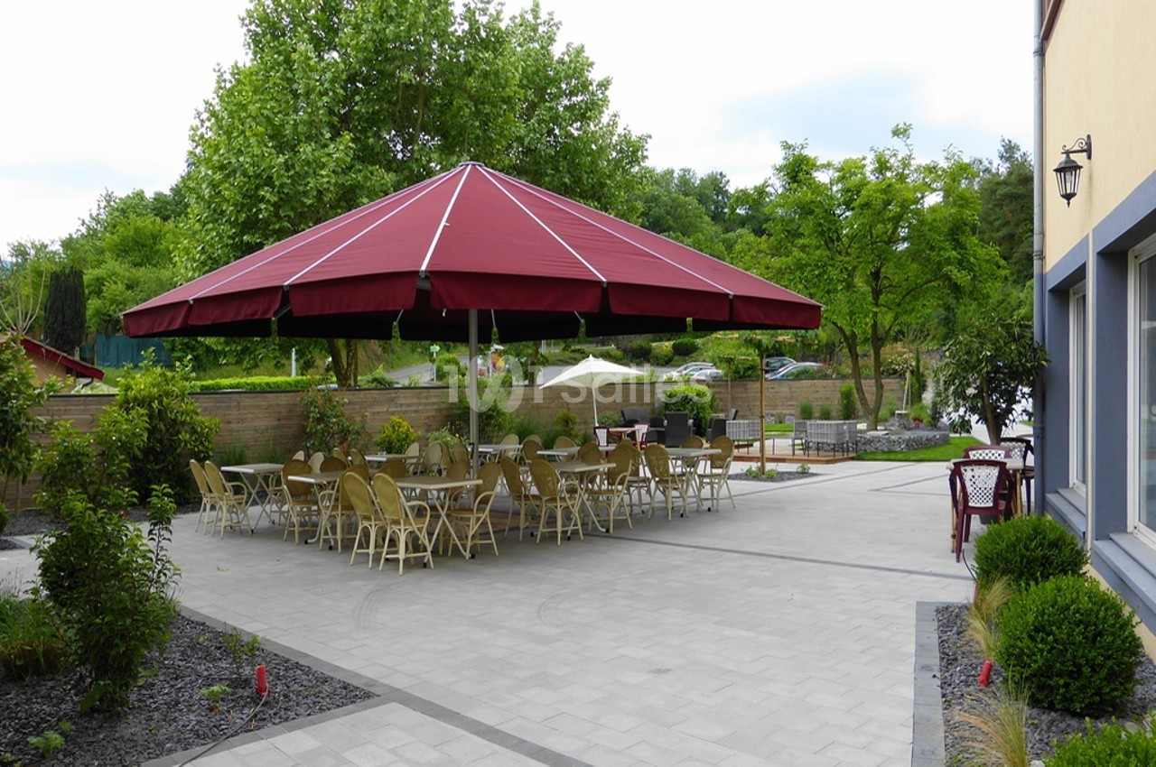 Terrasse extérieure avec tables et chaises sous un grand parasol rouge, entourée de verdure et de murs en pierre.