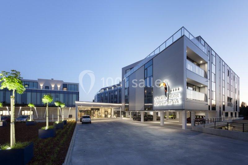 Façade moderne d'un hôtel avec enseigne lumineuse, parking et arbres décoratifs, photographiée au crépuscule.