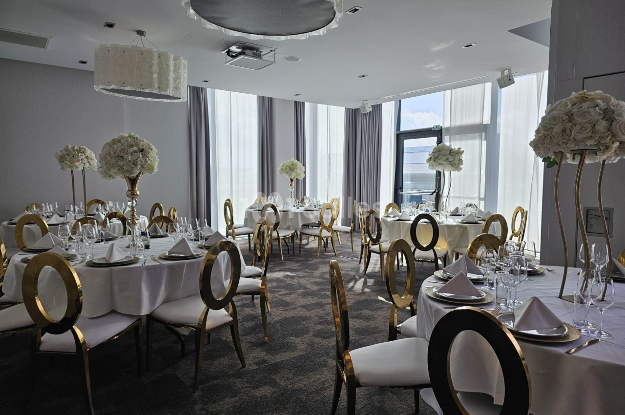 Salle de réception élégante avec tables rondes dressées, chaises dorées et décorations florales blanches.