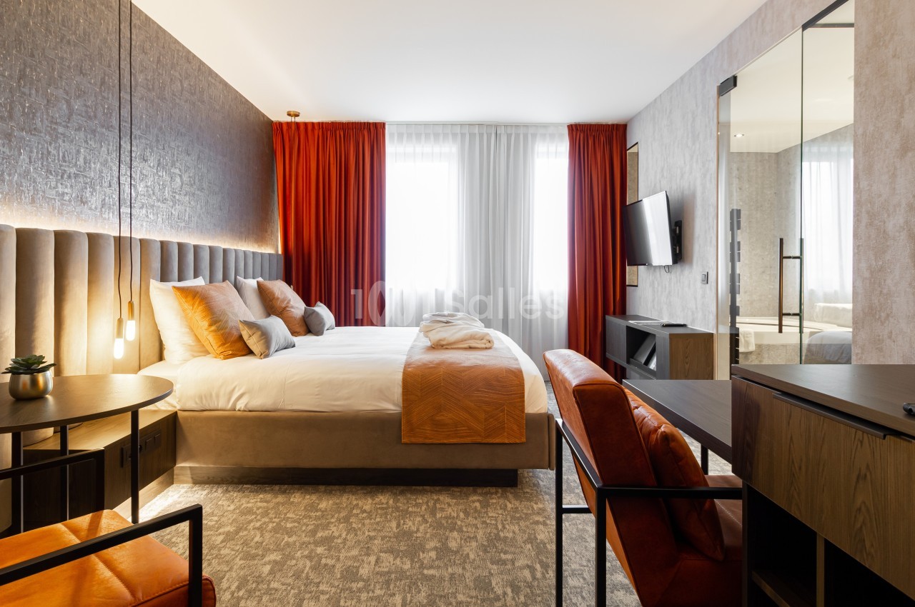 Chambre d'hôtel moderne avec lit double, fauteuils en cuir, bureau, télévision murale et grandes fenêtres avec rideaux.