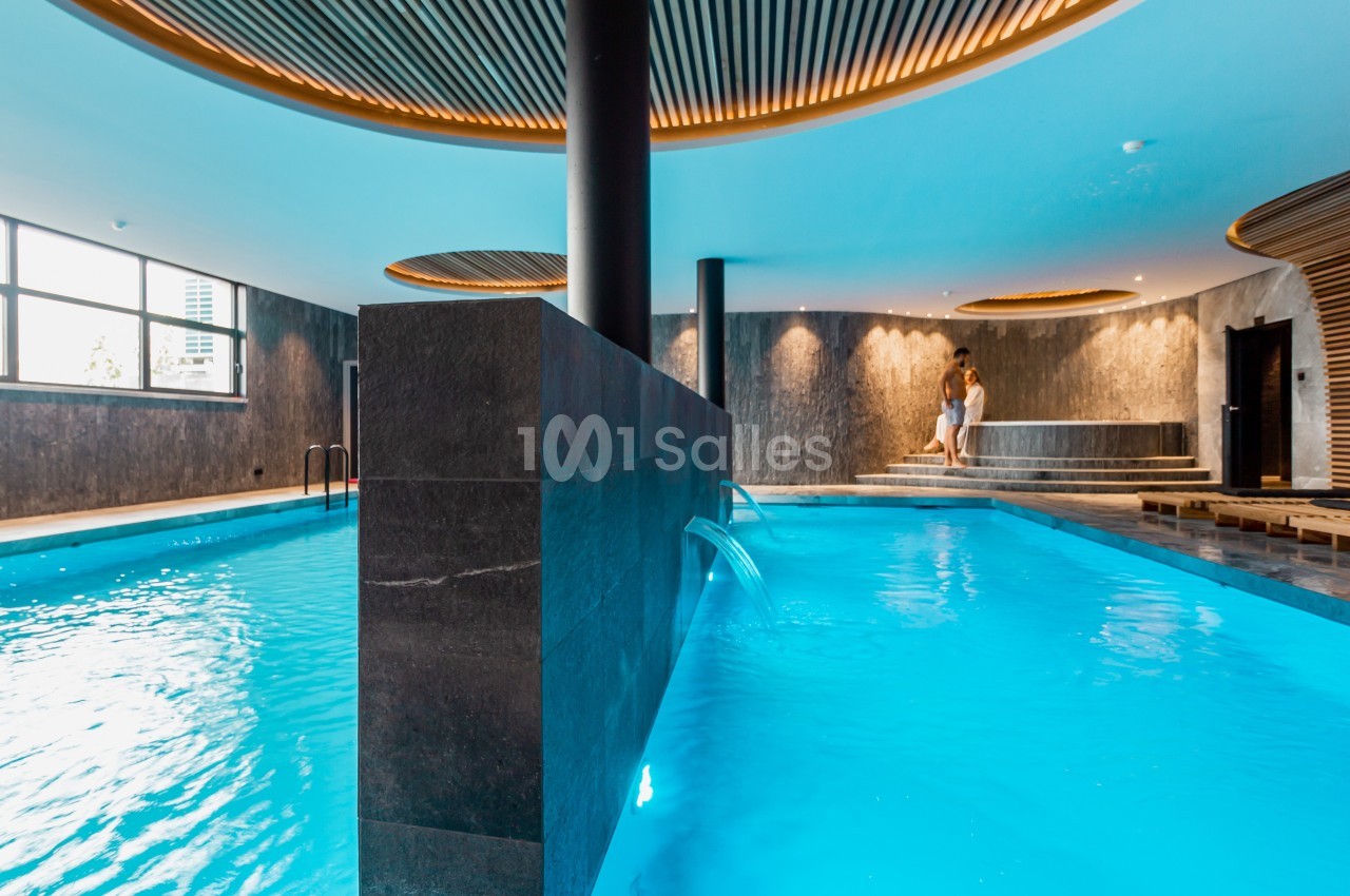 Piscine intérieure moderne avec éclairage tamisé, jets d'eau et espace détente en arrière-plan.