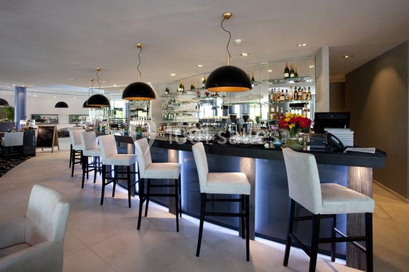 Bar moderne avec comptoir éclairé, tabourets rembourrés, suspensions noires et étagères garnies de bouteilles.