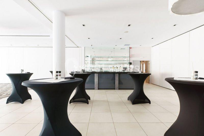 Salle lumineuse avec tables hautes noires et un bar moderne en arrière-plan.