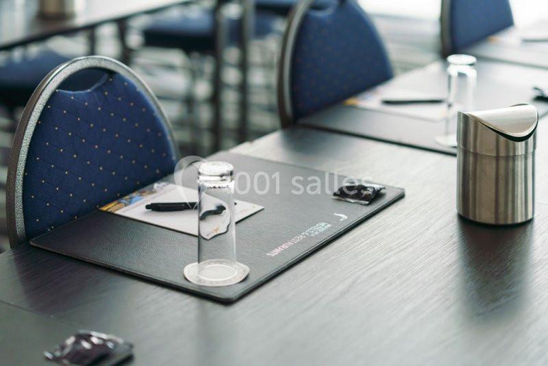 Salle de réunion avec des chaises bleues, des tables noires, des sous-mains, des stylos et des verres disposés.