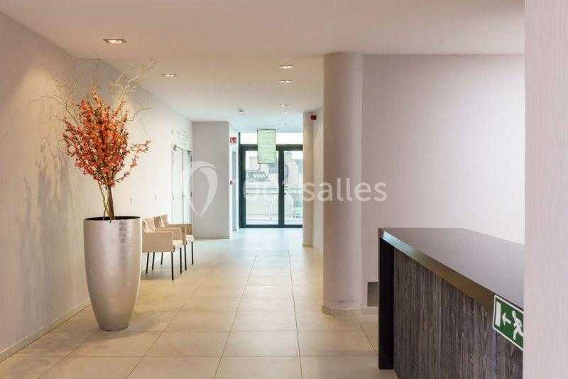 Un hall lumineux avec un comptoir sombre, un grand vase de fleurs orange et des chaises près d'une porte vitrée.