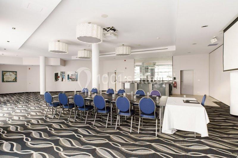 Salle de réunion lumineuse avec des chaises bleues autour de tables noires, un bar en arrière-plan et un écran blanc.