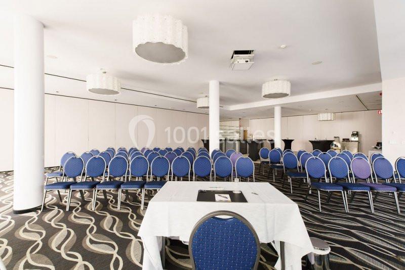 Salle de conférence lumineuse avec rangées de chaises bleues, table à l'avant et décor sobre.