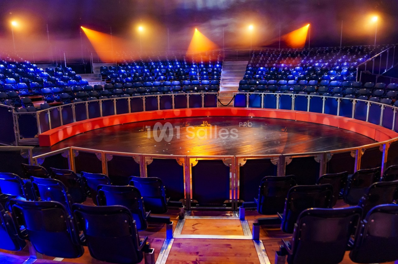 Gradins vides disposés en cercle autour d'une scène en bois éclairée par des projecteurs dans une salle de spectacle.