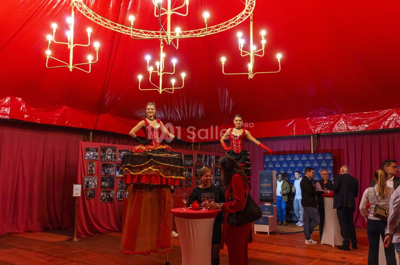 Deux artistes sur échasses en costumes rouges accueillent des invités dans une salle décorée de rideaux et lustres.