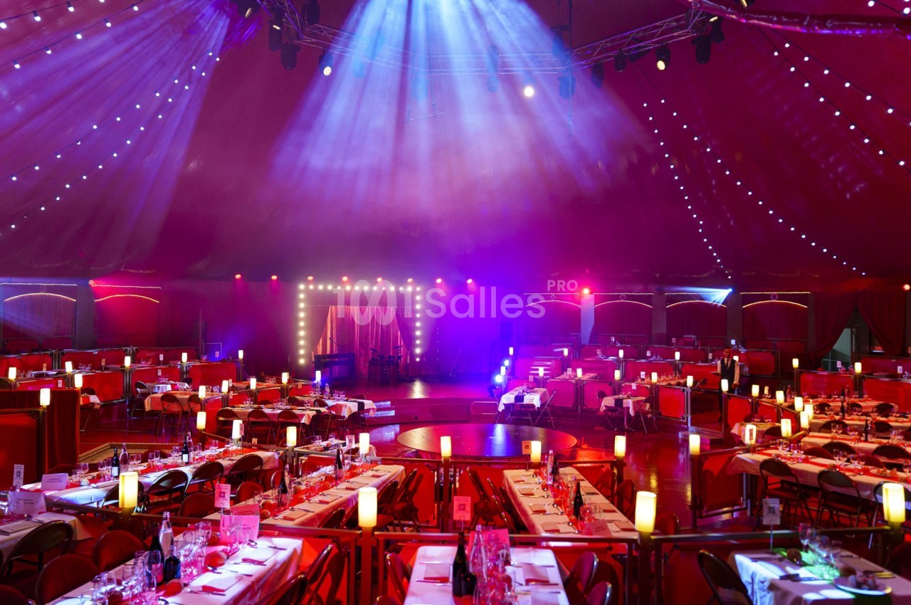 Salle de spectacle sous un chapiteau avec tables dressées, éclairages colorés et scène illuminée en arrière-plan.