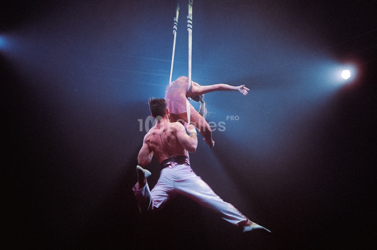 Deux artistes de cirque exécutent une figure acrobatique suspendus à des sangles sous un éclairage de scène.