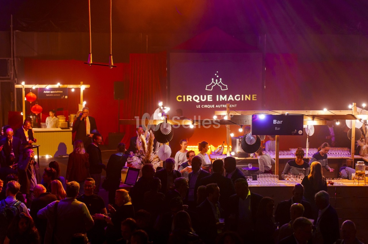 Foule rassemblée dans un espace éclairé avec des stands de restauration et une scène affichant ’Cirque Imagine’.