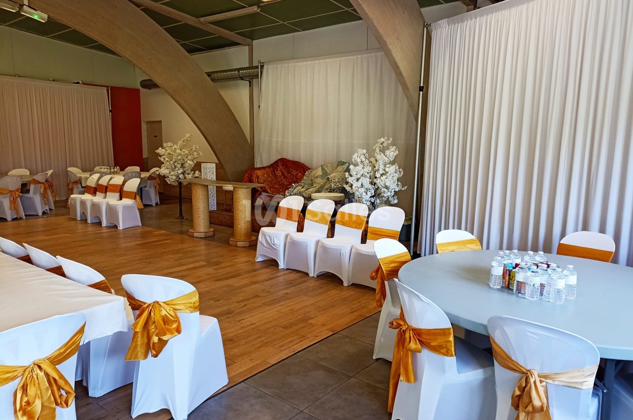 Salle décorée pour un événement avec des chaises blanches ornées de nœuds dorés, tables dressées et rideaux blancs.