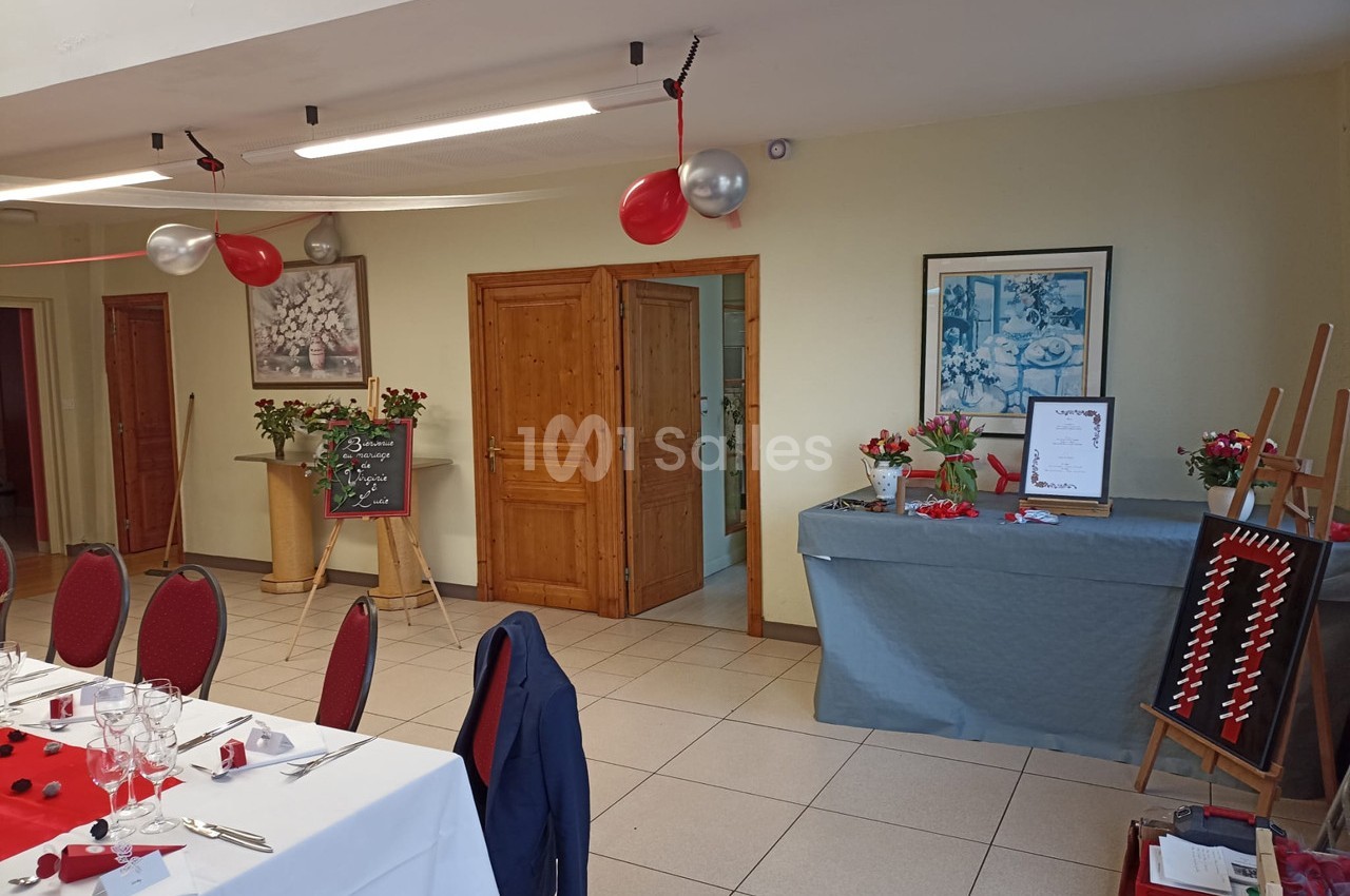 Salle décorée pour un événement avec tables dressées, ballons rouges, ardoise et table de présentation ornée de fleurs.