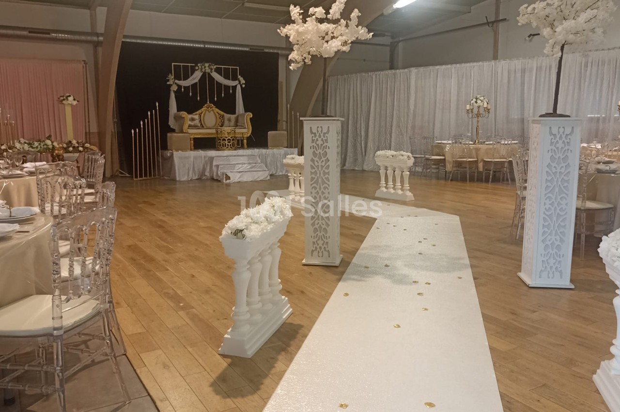 Salle décorée pour un mariage avec allée blanche ornée de fleurs, chaises transparentes et table d'honneur en arrière-plan.