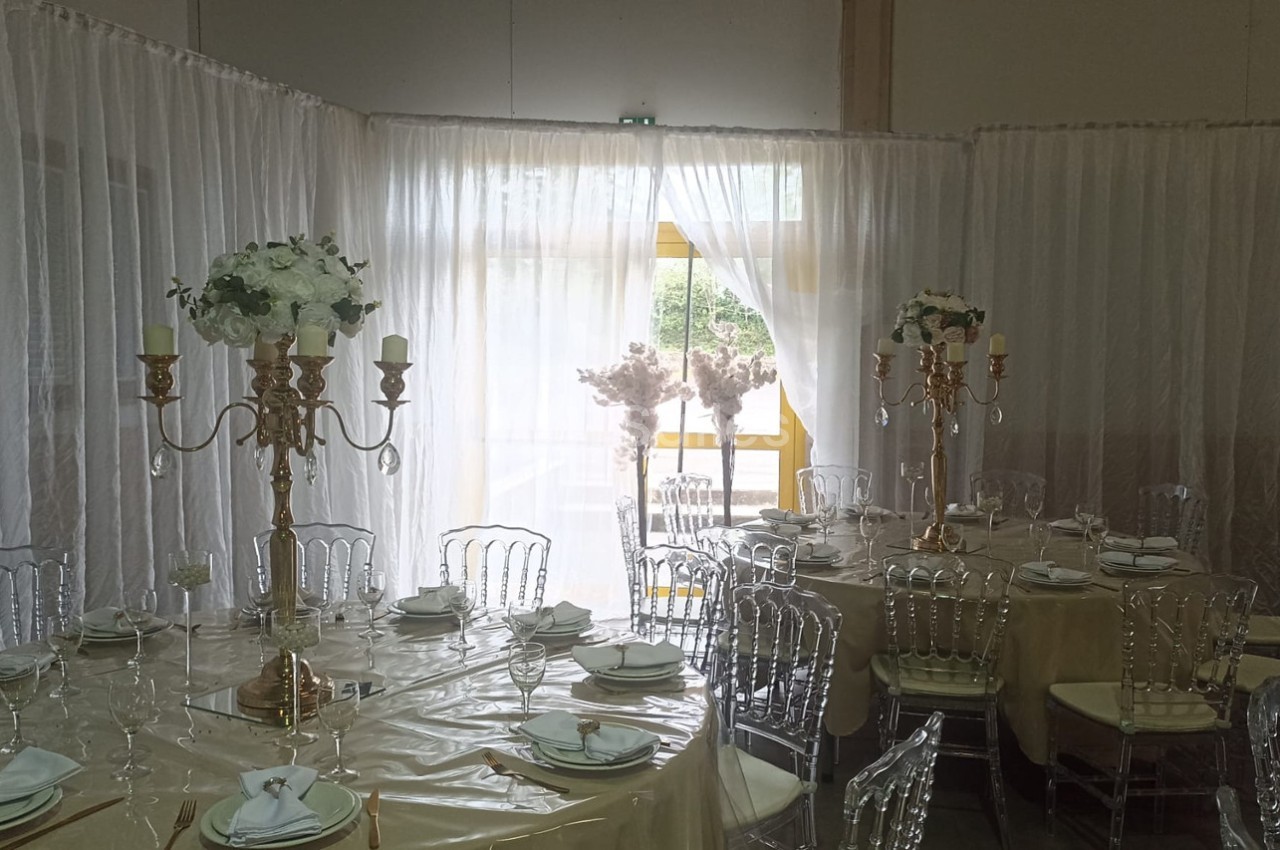 Salle décorée pour un événement avec tables rondes, nappes beiges, chaises transparentes et chandeliers dorés ornés de…