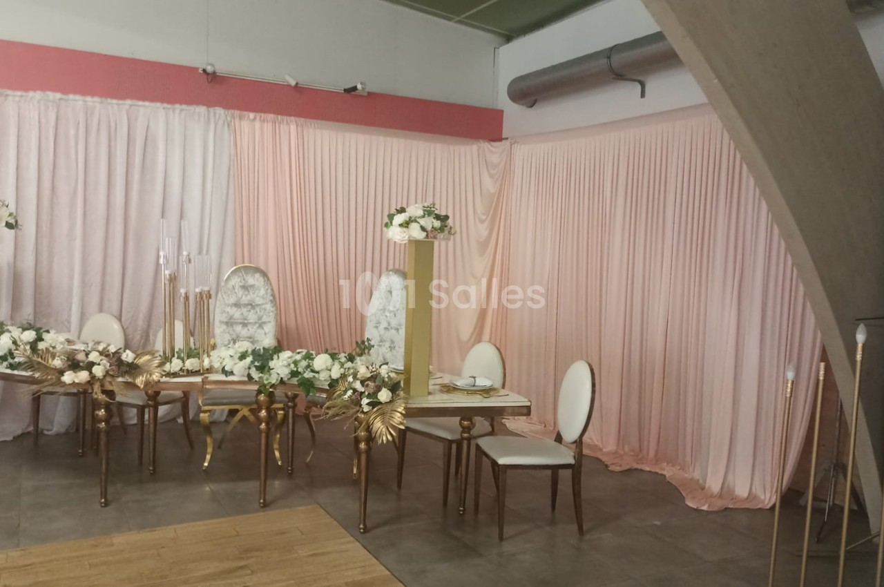 Salle décorée pour un événement avec des chaises blanches, une table dorée et des rideaux roses en arrière-plan.