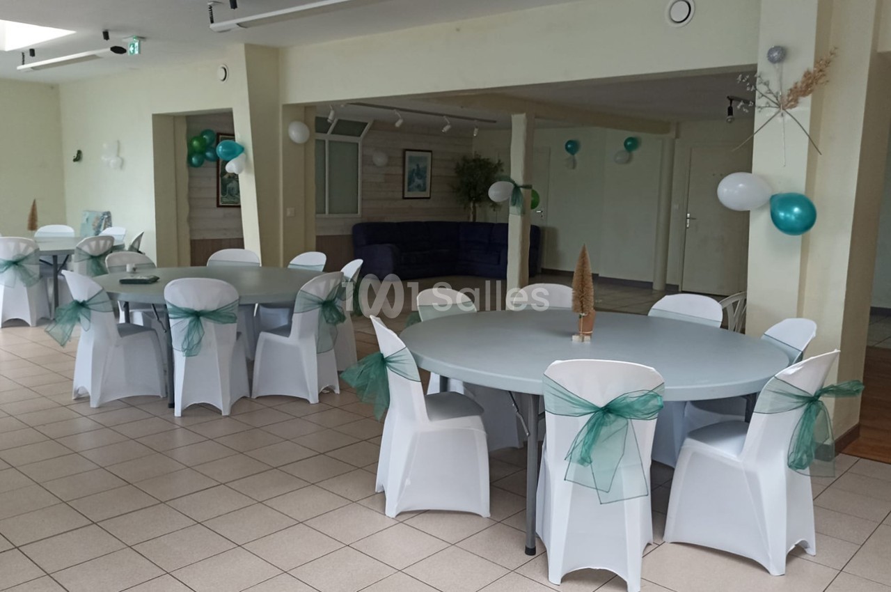 Salle décorée pour un événement avec des tables rondes, des chaises blanches ornées de rubans verts et des ballons.