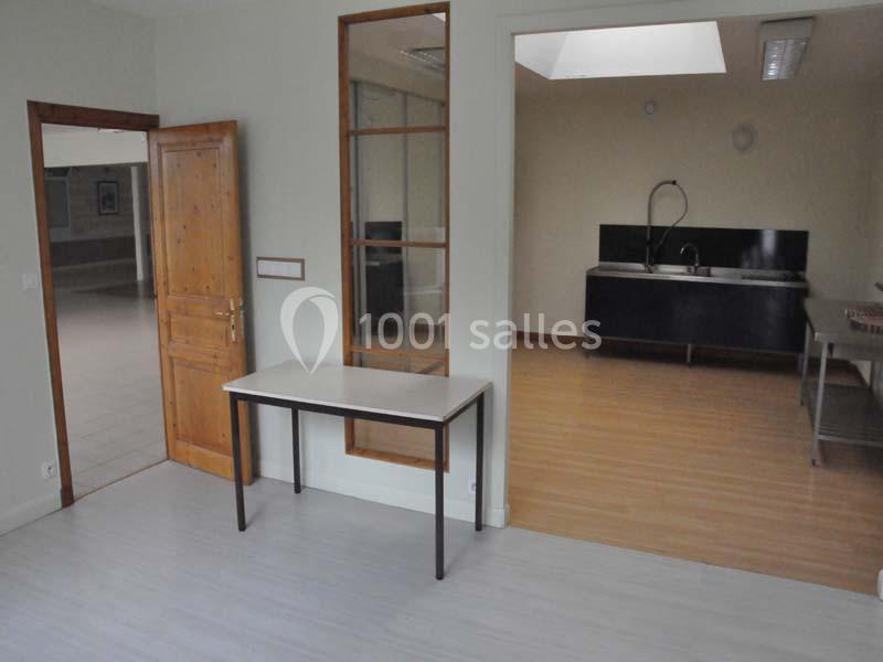 Salle avec une table simple, une porte ouverte menant à une autre pièce et une cuisine équipée visible au fond.