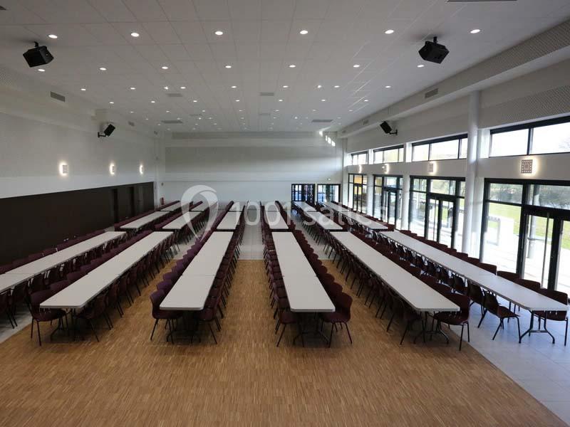 Salle spacieuse avec de longues tables alignées et des chaises, éclairée par de grandes baies vitrées.