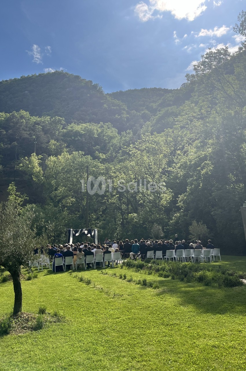 Cérémonie en plein air avec des invités assis, entourée de verdure et de collines sous un ciel ensoleillé.