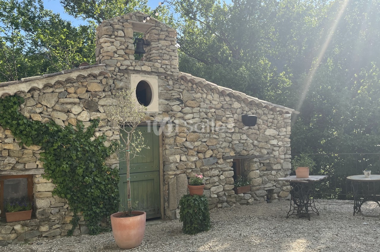 Petite maison en pierre entourée de végétation, avec une porte verte, des plantes en pot et une table en extérieur.