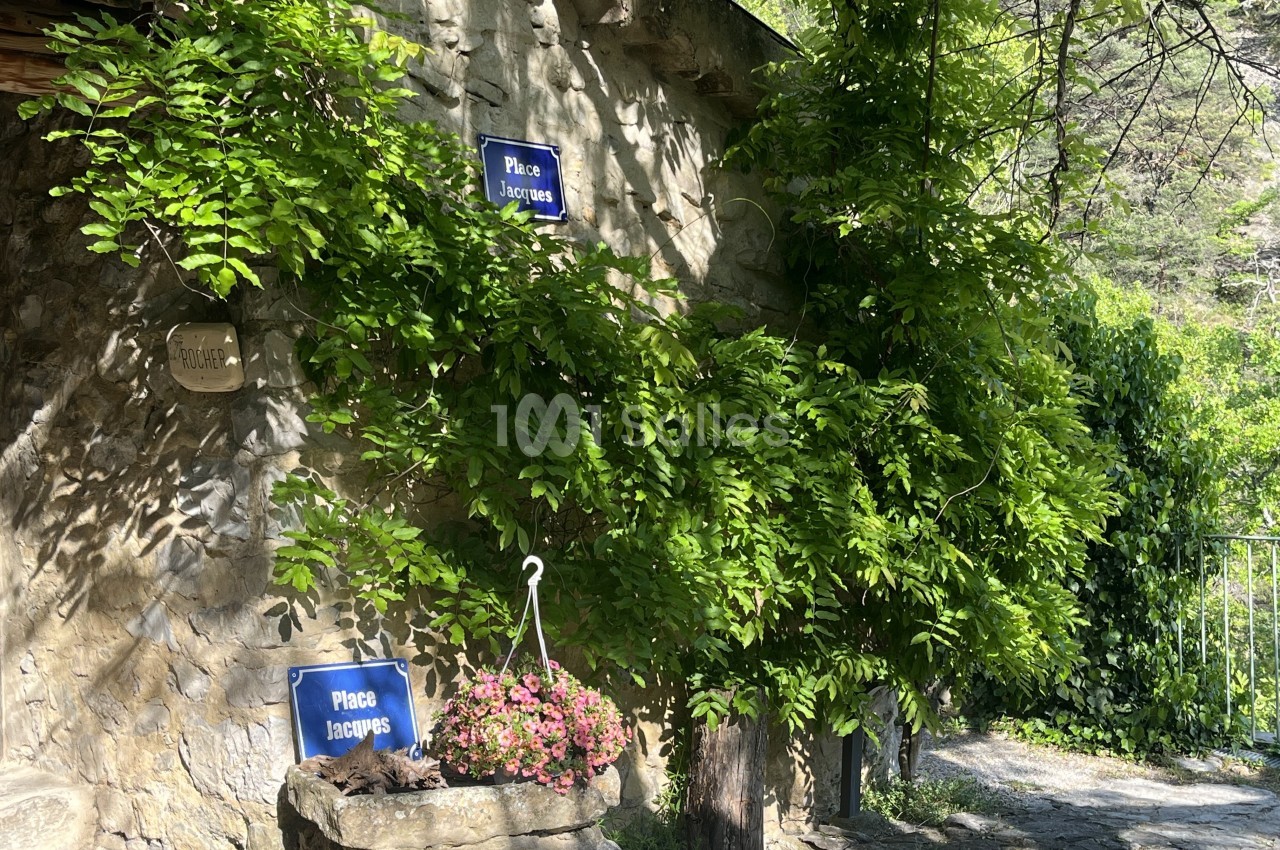 Façade en pierre avec des plantes grimpantes, deux panneaux ’Place Jacques’ et une jardinière fleurie au premier plan.