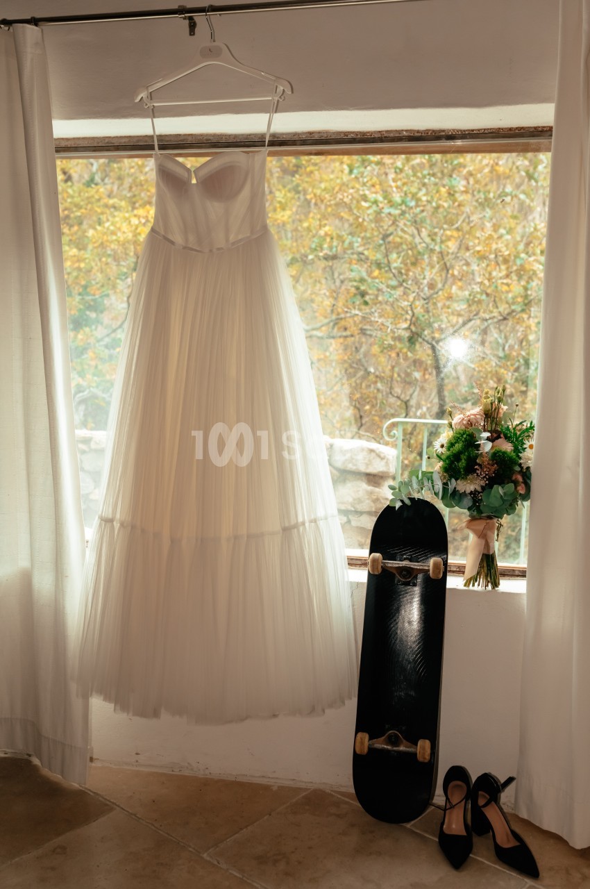 Robe de mariée suspendue près d'une fenêtre, avec un bouquet, un skateboard et des chaussures à talons posés au sol.