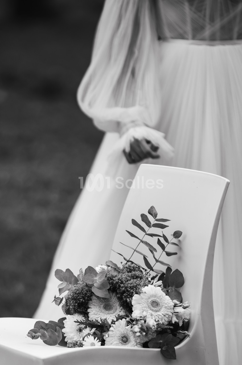 Une chaise blanche avec un bouquet de fleurs posé dessus, en arrière-plan une personne en robe claire floue.