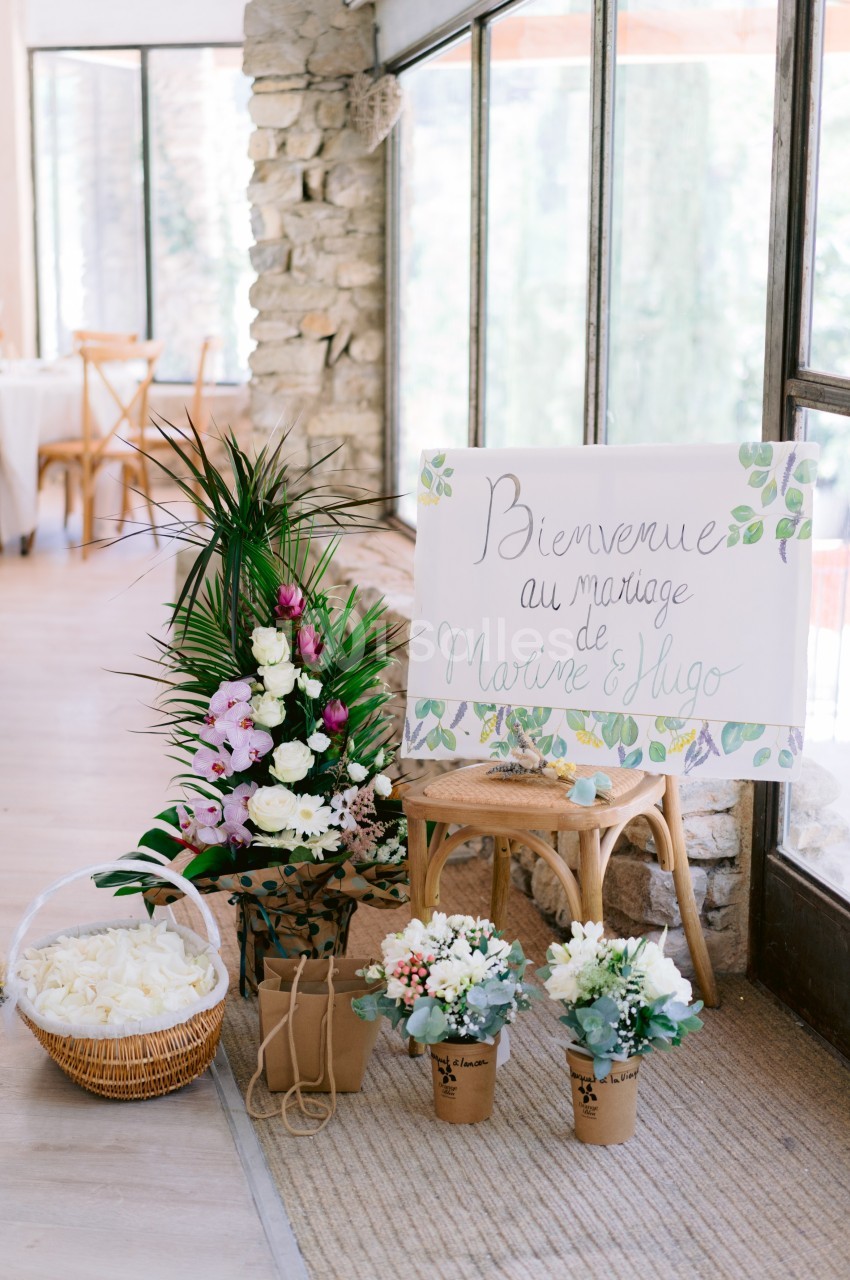 Panneau de bienvenue pour un mariage entouré de fleurs, paniers et plantes dans une salle lumineuse.