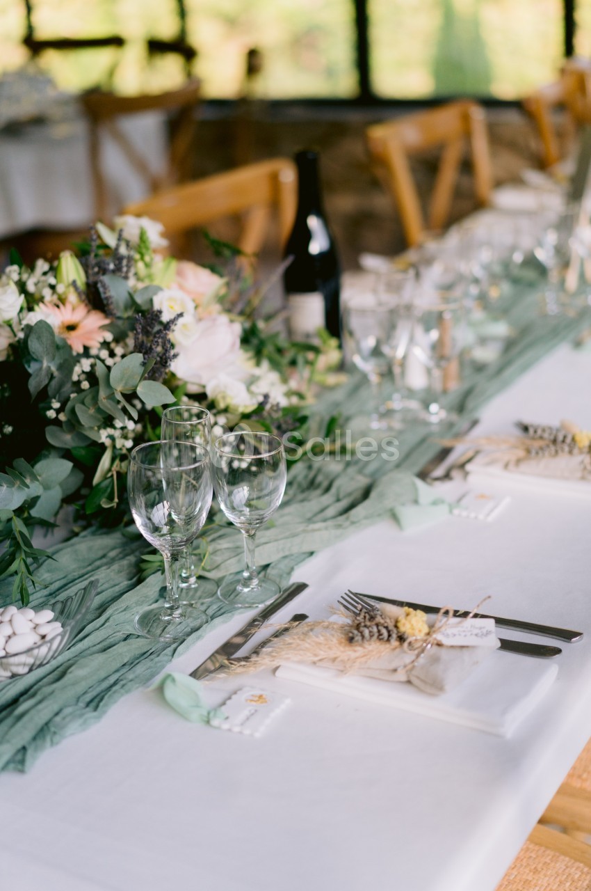 Table décorée pour un repas avec nappes blanches, verres, couverts, fleurs et éléments naturels.