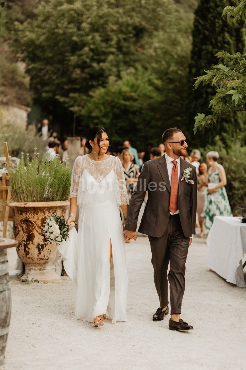 Un couple habillé pour un mariage marche main dans la main dans un jardin extérieur, entouré d'invités.