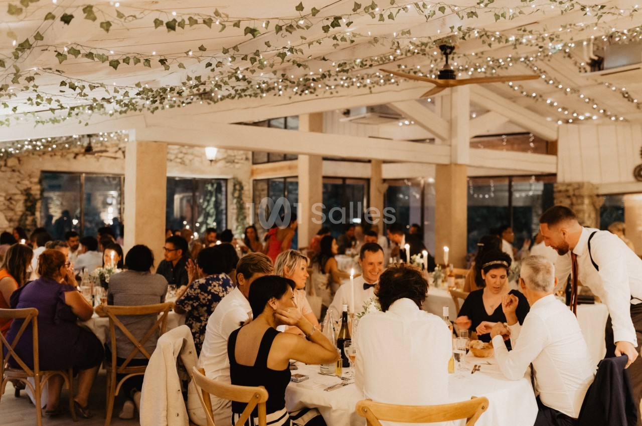 Salle de réception lumineuse avec des invités dînant à des tables décorées, sous des guirlandes lumineuses et des plantes…