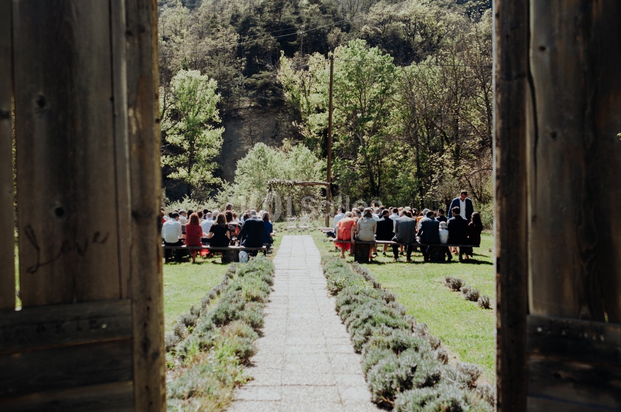 Vue d'une cérémonie en plein air avec des invités assis, entourée de verdure et encadrée par des portes en bois ouvertes.