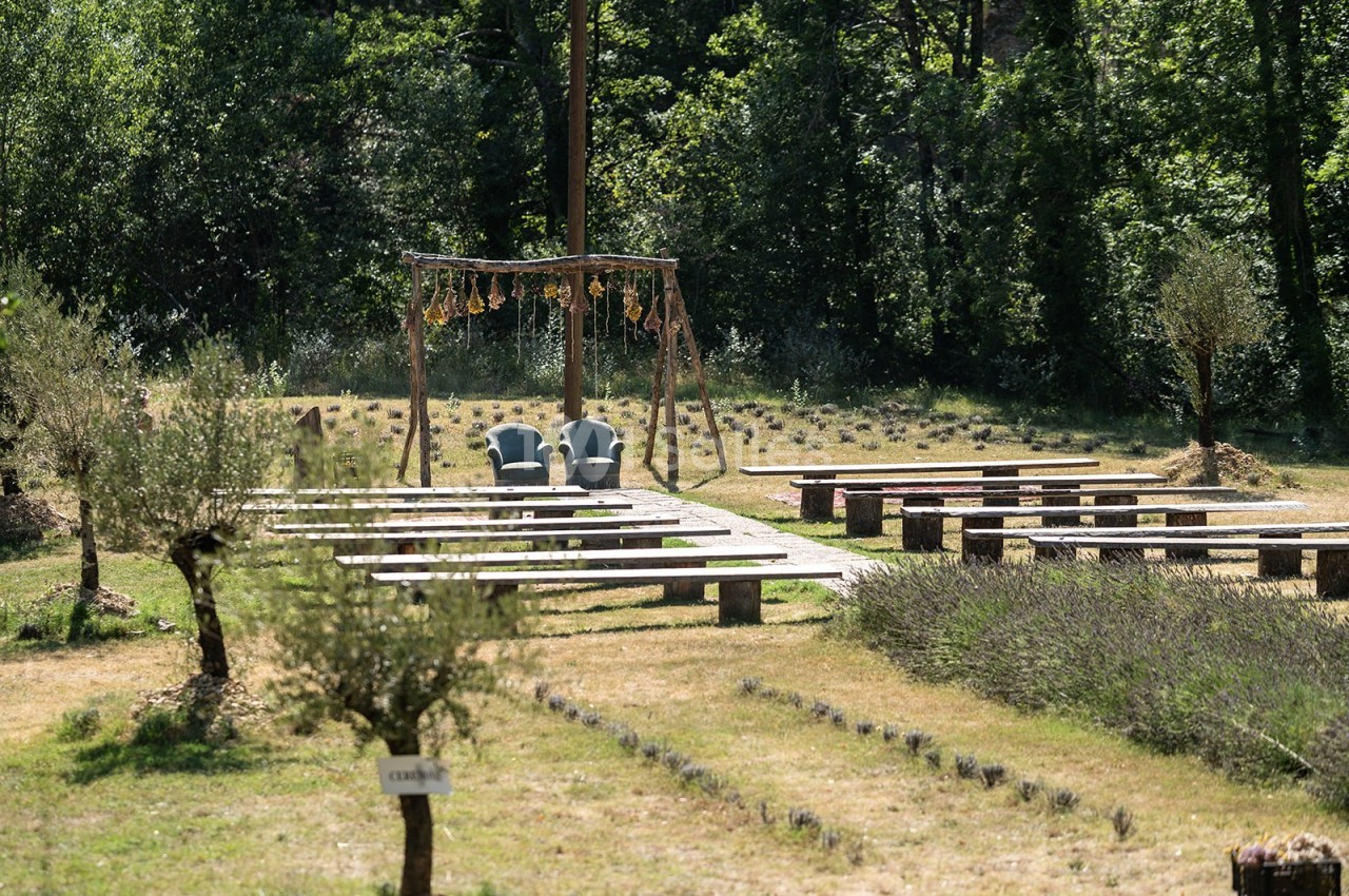 Chaises disposées sous une arche décorée, entourées de rangées de bancs en bois dans un cadre naturel verdoyant.