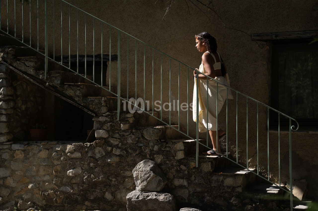 Une femme monte un escalier en pierre à l'extérieur d'une maison, éclairée par une lumière naturelle.