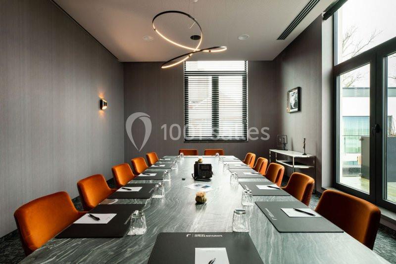 Salle de réunion moderne avec table en marbre, chaises orange, lumière suspendue et fenêtres avec stores.