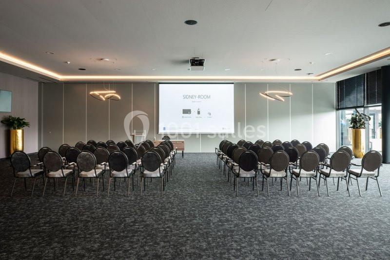 Salle de conférence moderne avec rangées de chaises alignées face à un écran de projection allumé.