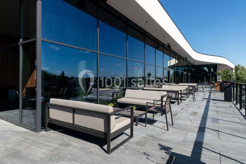 Terrasse moderne avec des canapés et tables alignés le long d'une façade vitrée, sous un ciel dégagé.