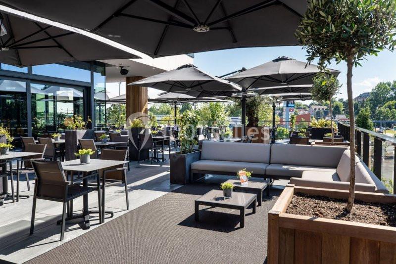 Terrasse moderne avec tables, chaises, canapés et parasols, entourée de plantes et baignée de lumière naturelle.