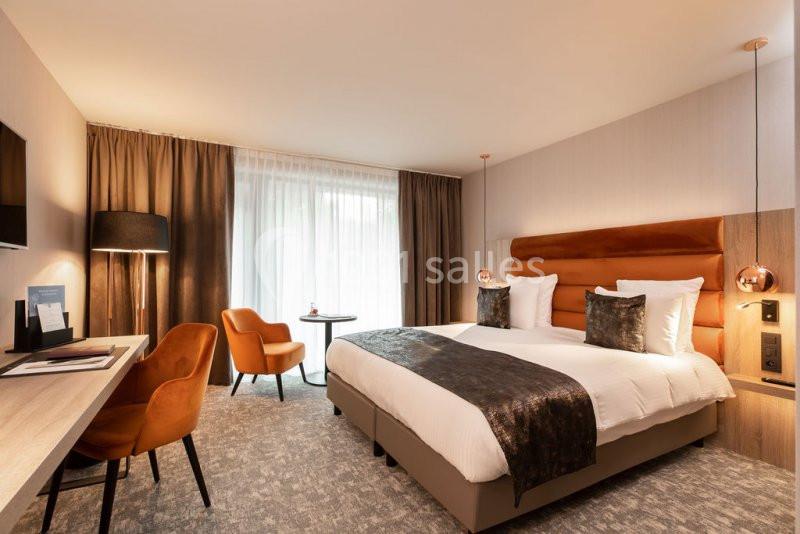 Chambre d'hôtel moderne avec lit double, bureau, fauteuils orange, lampadaire et grande fenêtre avec rideaux.